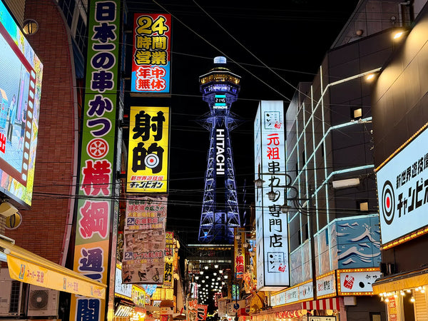 Beleuchtete Straße im Shinsekai-Viertel in Osaka mit bunten Leuchtreklamen und dem Tsutenkaku-Turm bei Nacht – Symbol für Japans modernes Stadtleben